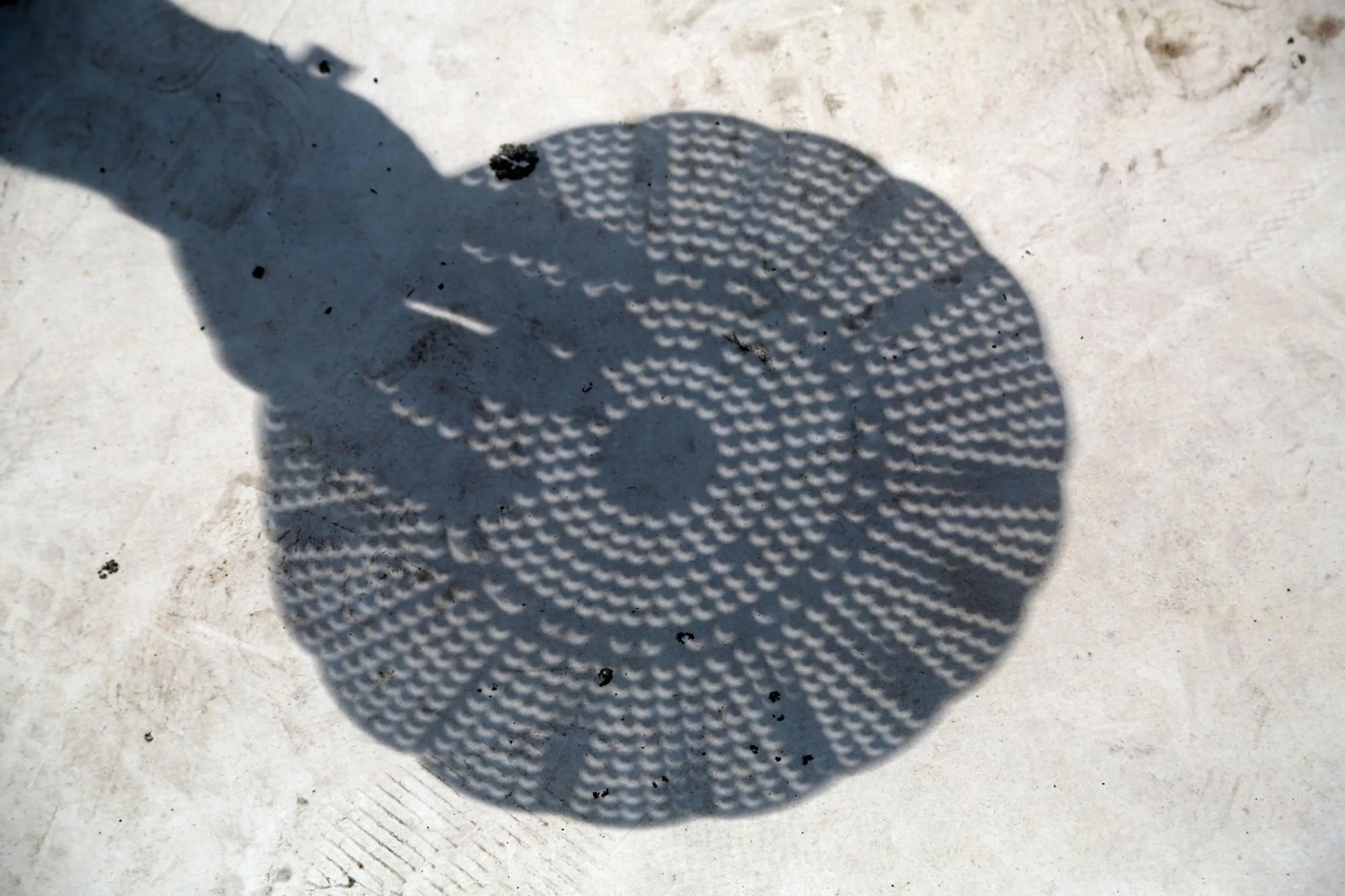 Imagen que muestra cómo los agujeros circulares de un colador proyectan figuras de media luna en el suelo durante las fases parciales de un eclipse solar.