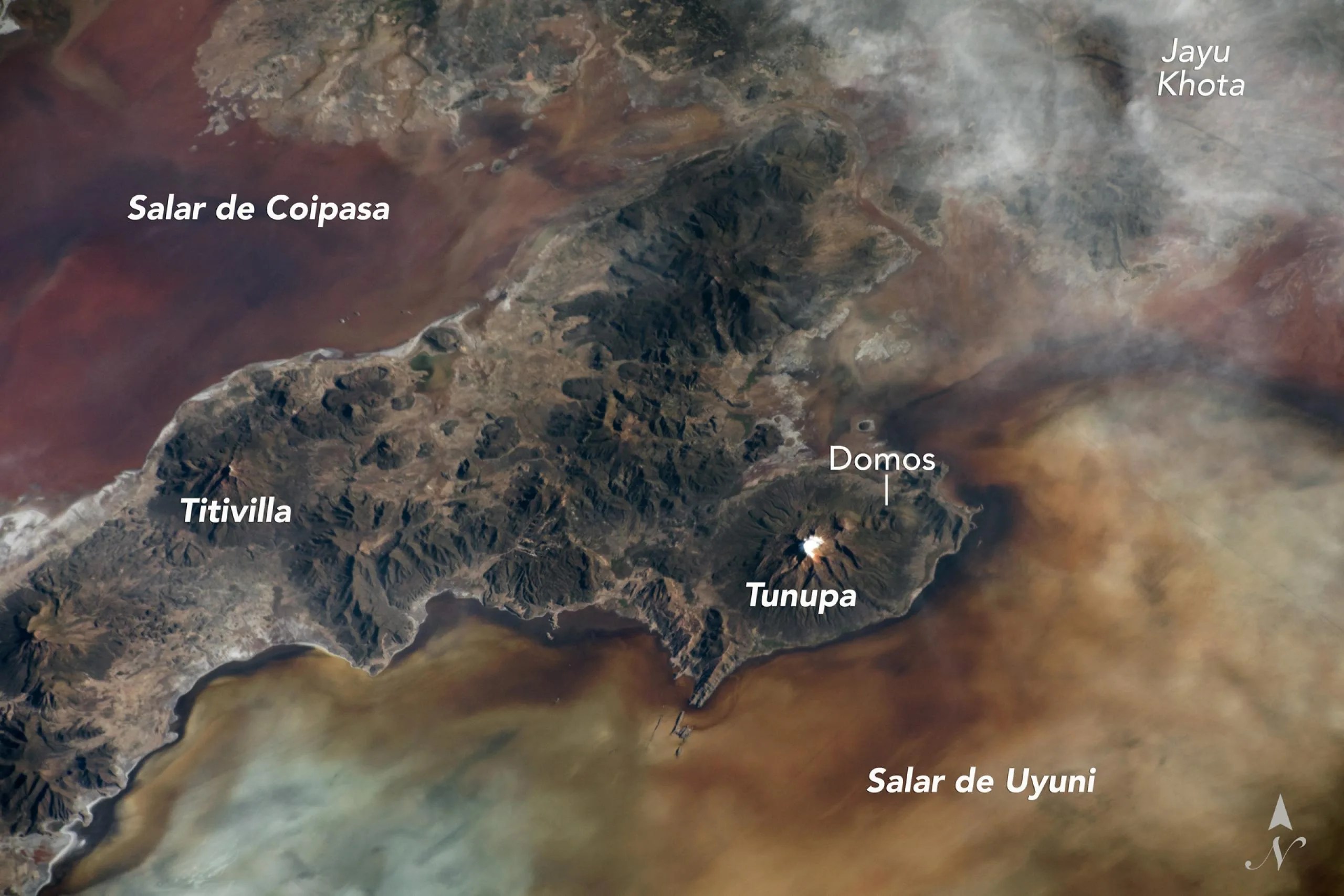Imagen del volcán Tunupa en Bolivia tomada desde el espacio. La fotografía muestra al volcán situado en una península entre dos de las salinas más grandes de Bolivia, el Salar de Uyuni y el Salar de Coipasa. Estas salinas tienen una cobertura de sedimentos y poblaciones microbianas variables, lo que produce áreas con tonos más oscuros y más claros en la superficie. Se observan los flancos del cono del volcán cortados por valles que fueron erosionados por antiguos glaciares y flujos de corrientes. Los domos