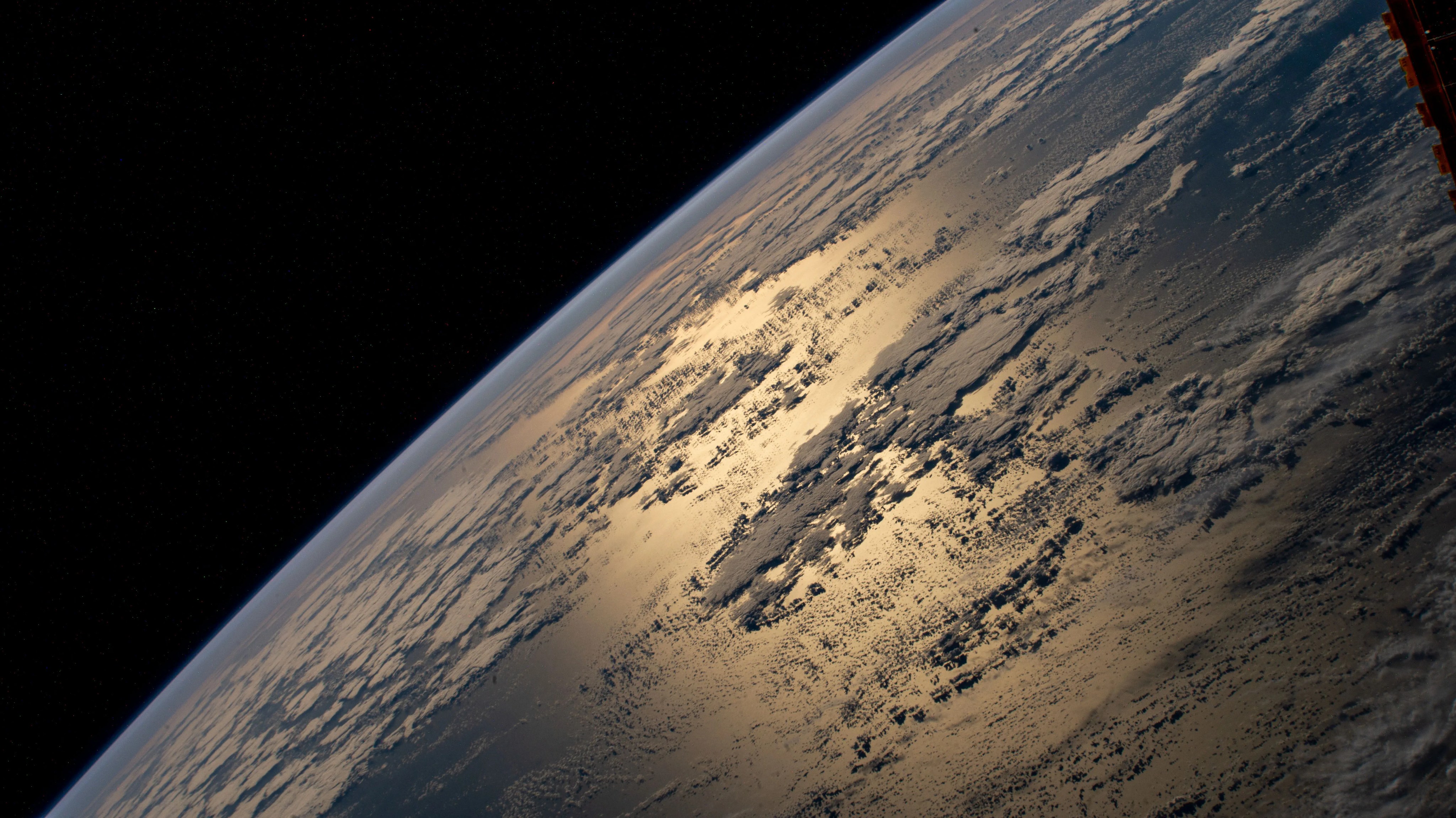 Imagen de la Tierra vista desde la Estación Espacial Internacional. La luz solar se refleja en el océano Atlántico. La Tierra, que ocupa tres cuartas partes de cuadro, muestra una capa de nubes rodeando un área donde se puede ver reflejada la luz solar en el océano.