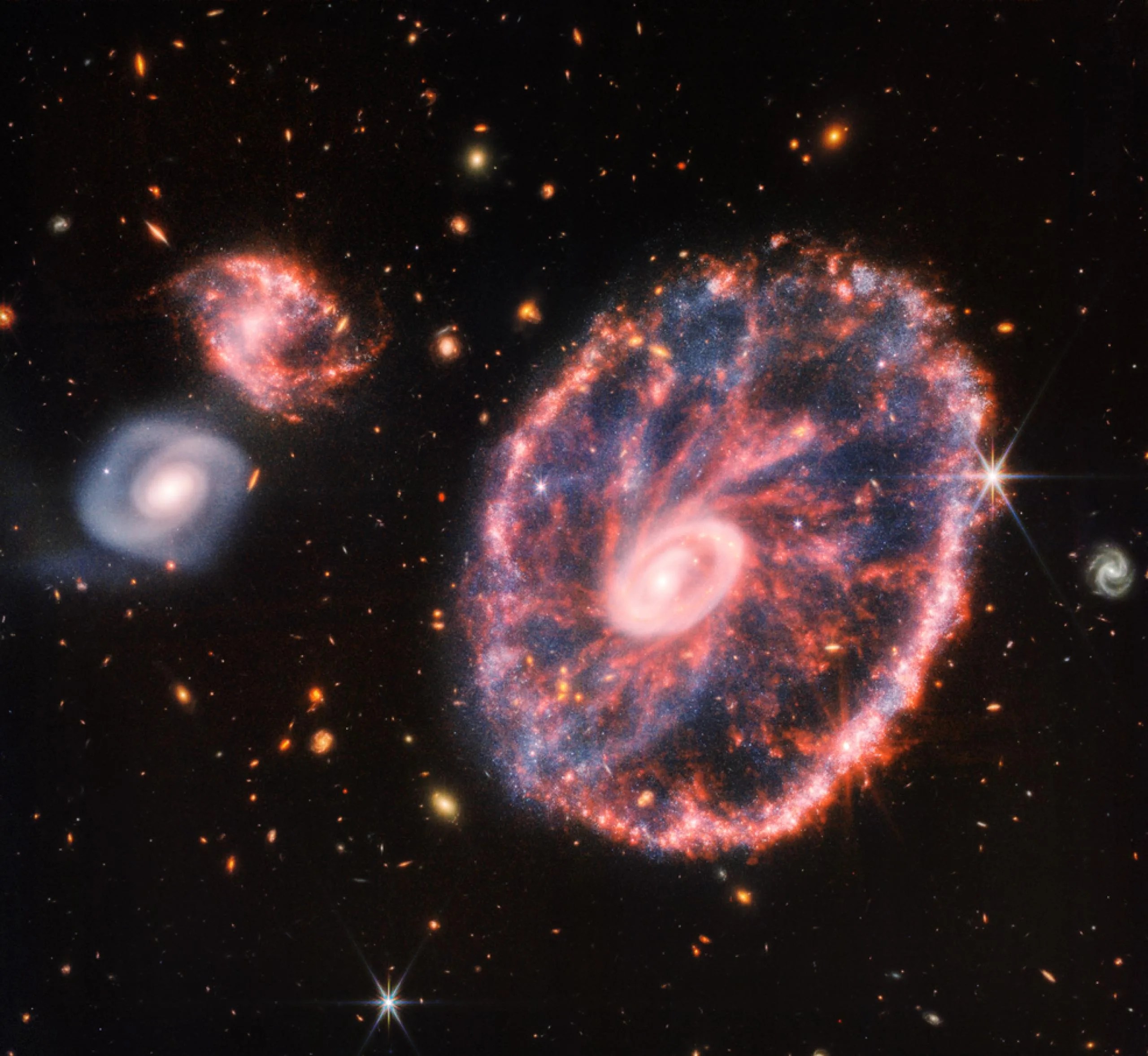 Una gran galaxia a la derecha, con dos galaxias compañeras mucho más pequeñas a la izquierda, a las 10 y a las 9 horas. La gran galaxia parece una rueda moteada, con un anillo exterior ovalado y un pequeño anillo interior que no está en el centro. El anillo exterior contiene penachos rosados como los radios de la rueda, con regiones azules polvorientas entre ellos. Las zonas rosas son polvo de silicatos, mientras que las azules son focos de estrellas jóvenes y polvo de hidrocarburos. El anillo interior es m