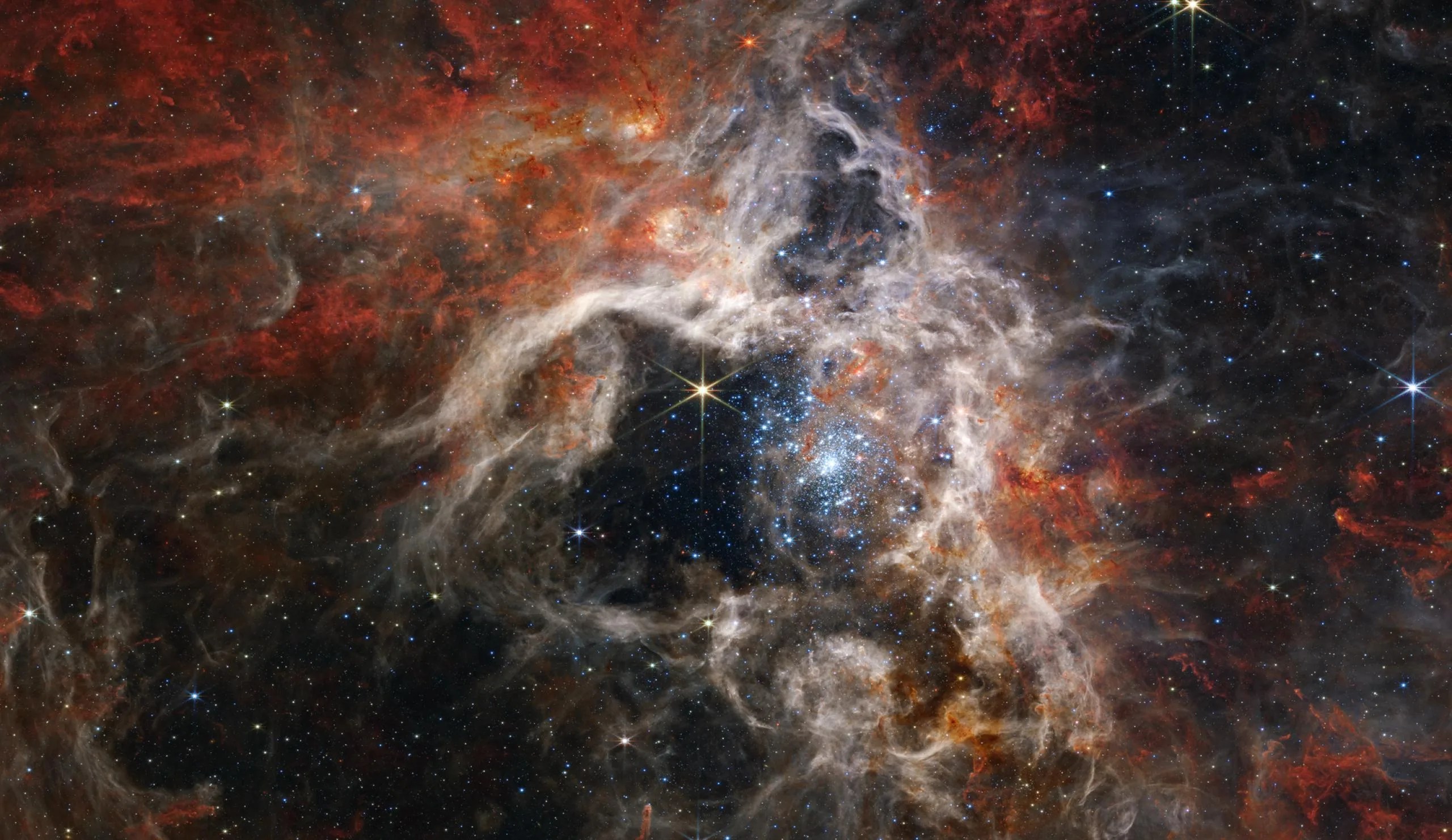 Una imagen espacial captada por el telescopio Webb. Esponjosas nubes de una nebulosa, de color tostado con reflejos parduzcos, rodean una zona central negra. Dentro de esa zona, el punto focal de la imagen es una gran estrella amarilla con ocho puntas largas y delgadas. A la derecha de la estrella está un brillante cúmulo de estrellas de forma ovalada. Las estrellas dentro del cúmulo aparecen como pequeños destellos de color azul pálido. El cúmulo está más densamente poblado en su núcleo y se dispersa hacia