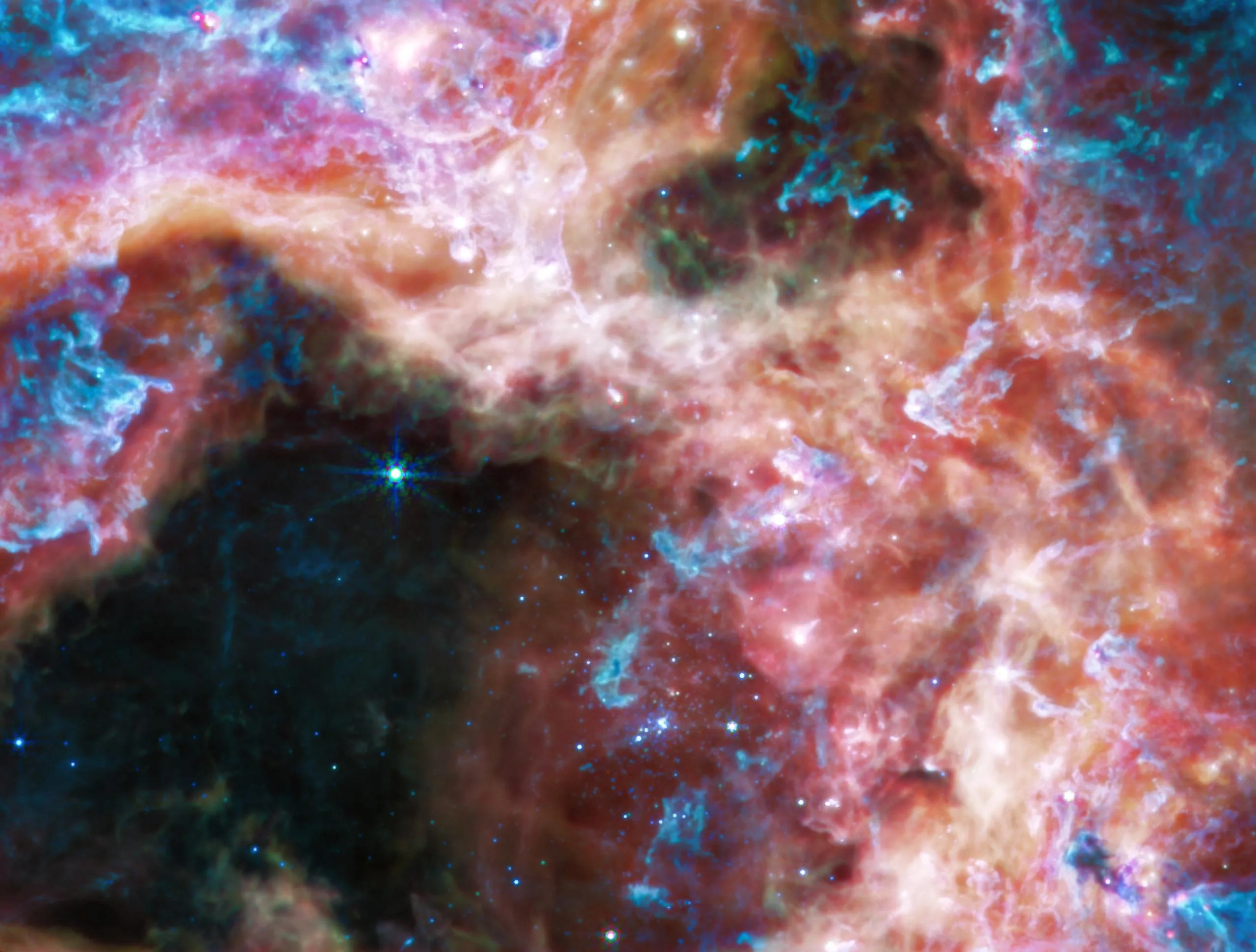 En las longitudes de onda de luz más largas captadas por su instrumento de infrarrojo medio (MIRI, por sus siglas en inglés), Webb se enfoca en el área que rodea el cúmulo estelar central y revela una vista muy diferente de la nebulosa de la Tarántula. En esta luz, se desvanece el brillo de las jóvenes estrellas calientes del cúmulo y aparece el resplandor del gas y el polvo. Abundantes hidrocarburos iluminan la superficie de las nubes de polvo, que se muestran en azul y morado.