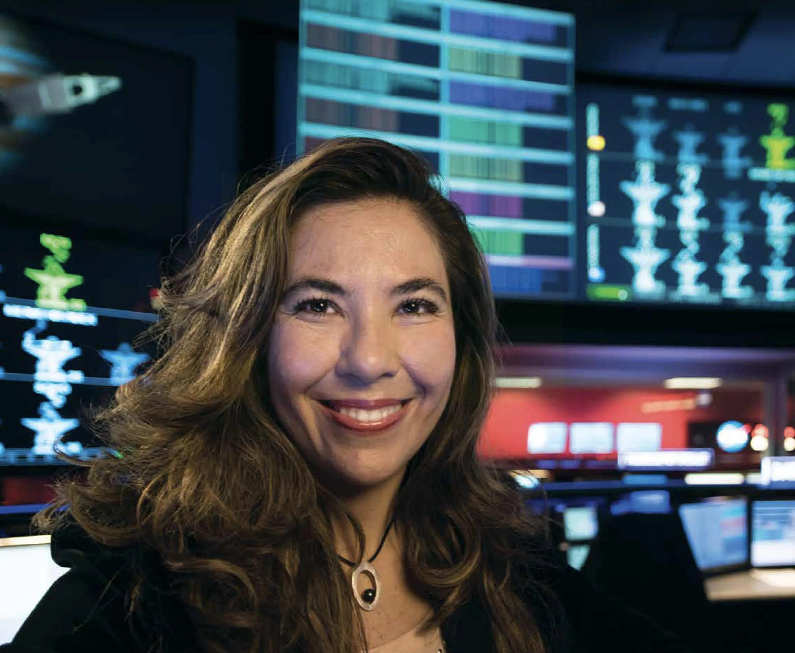 La Dra. Michela Muñoz Fernández, una mujer de pelo castaño claro y largo, sonríe a la cámara en primer plano. En el fondo se ven computadoras encendidas. Crédito de la imagen: NASA