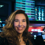 La Dra. Michela Muñoz Fernández, una mujer de pelo castaño claro y largo, sonríe a la cámara en primer plano. En el fondo se ven computadoras encendidas. Crédito de la imagen: NASA