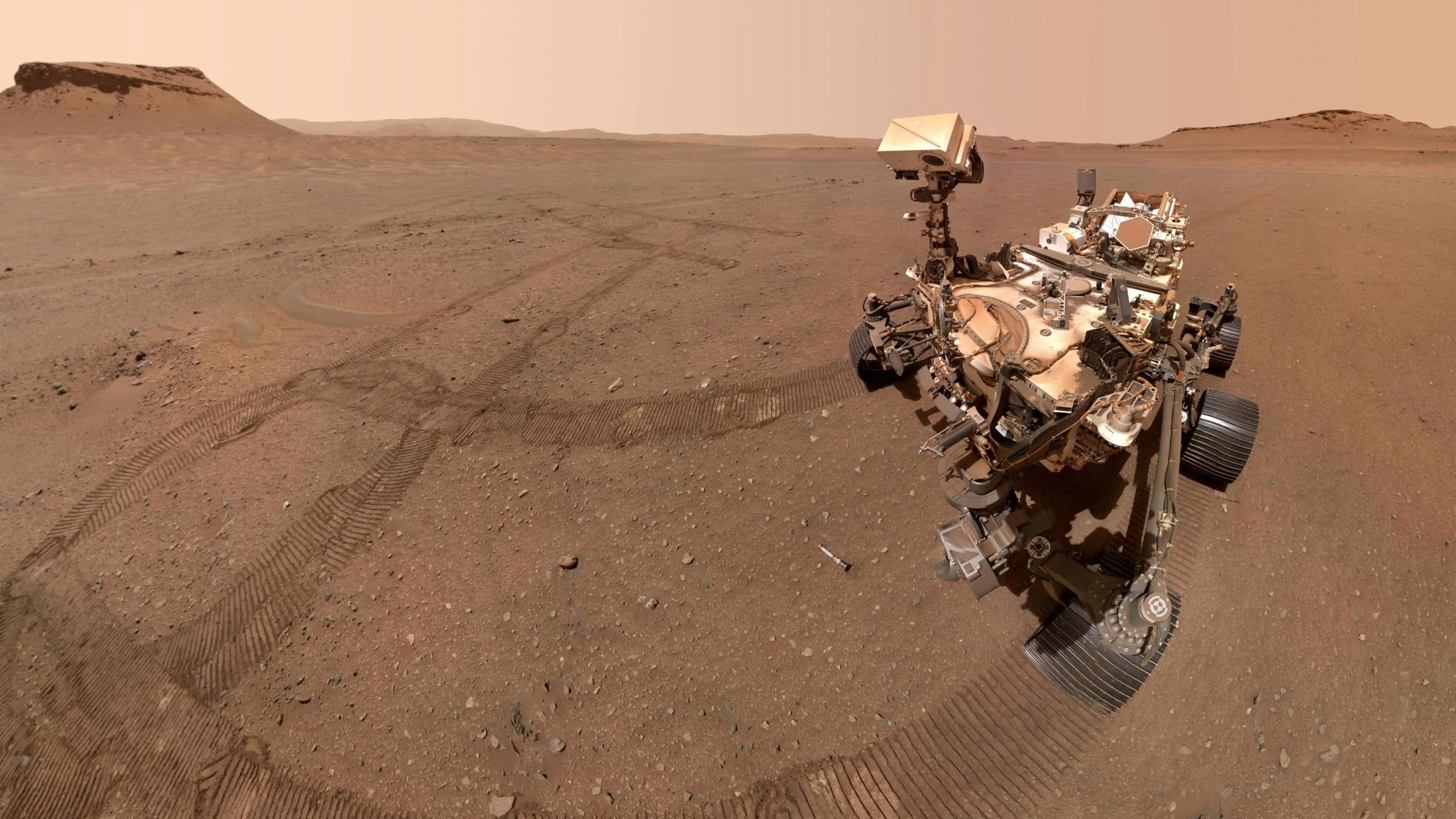 Esta imagen es una selfie sacada por el rover Perseverance de la NASA en Marte. Se ve parte del rover mirando hacia uno de los 10 tubos de muestras almacenados en el depósito que creó en un área apodada “Three Forks”. El fondo muestra el paisaje marciano, donde predomina el color rojizo.