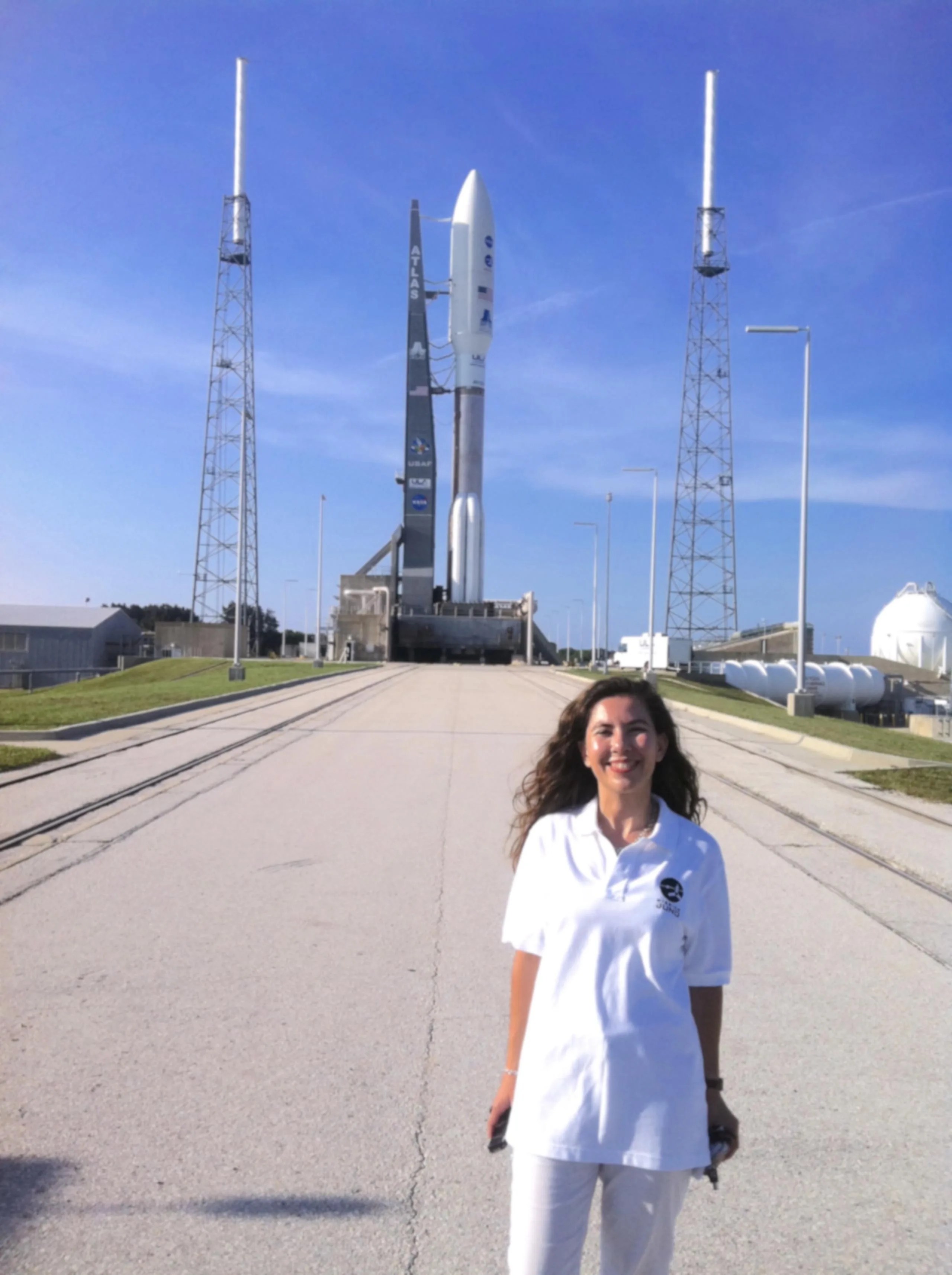La Dra. Michela Muñoz Fernández, una mujer de pelo largo y castaño, viste de blanco. Está parada mirando a la cámara y sonriendo, abajo a la derecha de la imagen. Al fondo, se ve un cohete blanco antes de despegar hacia el espacio con la nave espacial Juno. El cielo azul está mayormente despejado.