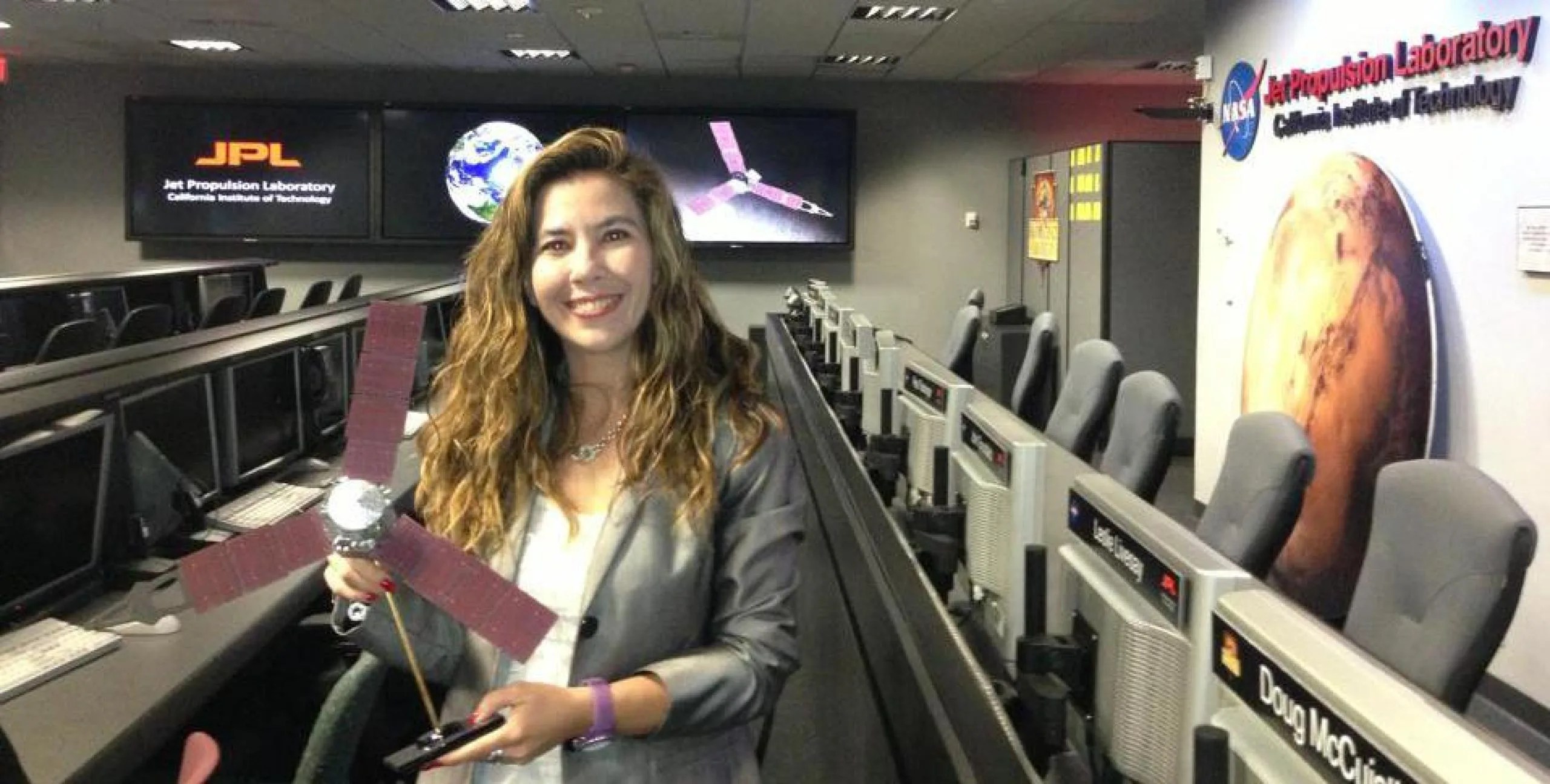 La Dra. Michela Muñoz Fernández aparece en primer plano vistiendo una chaqueta gris. Sonríe mirando a cámara mientras sostiene un modelo de la nave espacial Juno de la NASA. En el fondo se aprecia una sala de control en el Laboratorio de Propulsión a Chorro de la NASA (JPL).