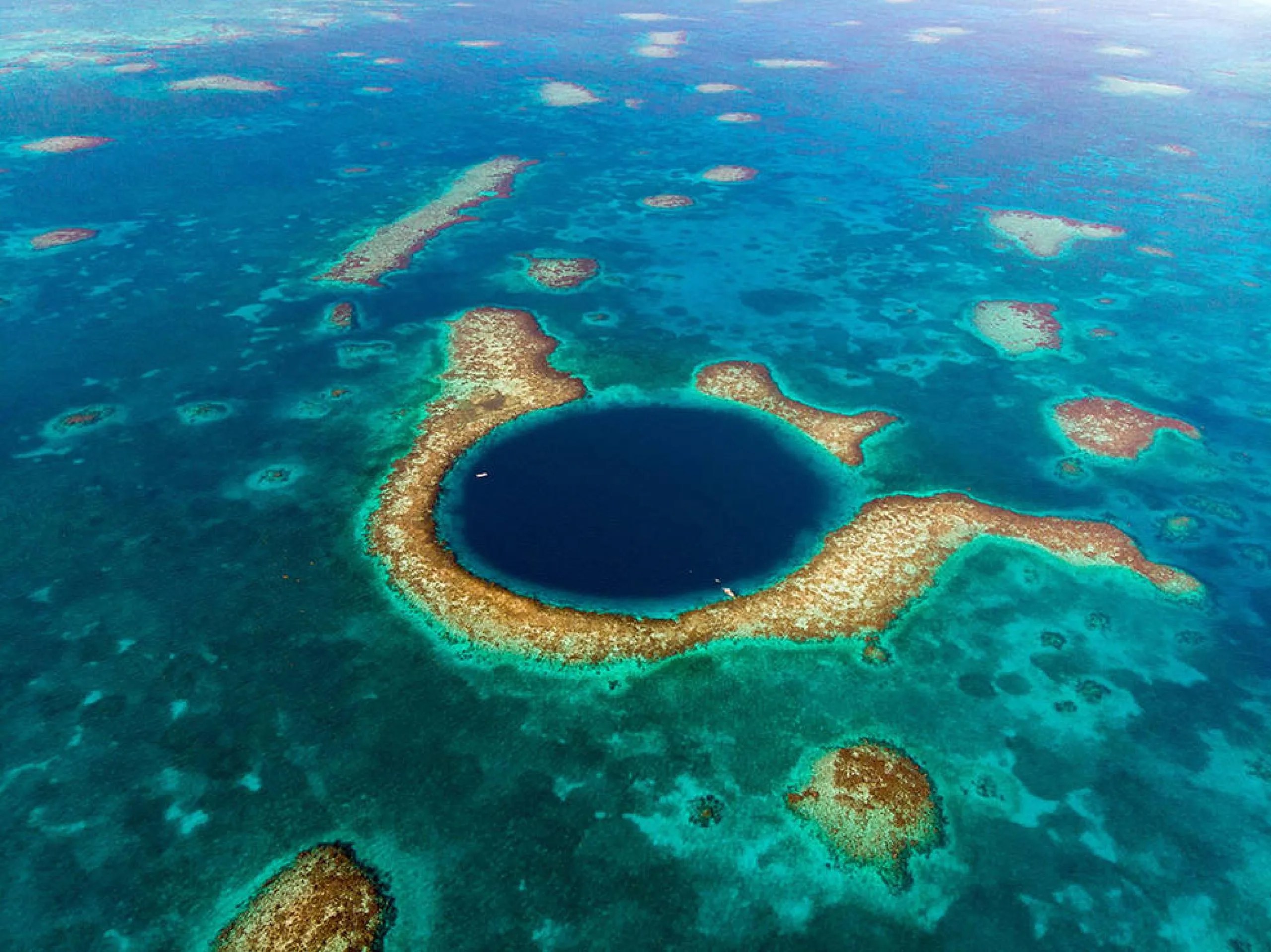 Foto aérea de una profunda cueva marina conocida como el Gran Agujero Azul, que forma parte del sistema de barrera de coral de Belice. Un círculo azul intenso en el centro de la imagen está rodeado por una sección de coral marrón claro. Más parches de coral marrón claro están dispersos por el agua de color turquesa circundante.