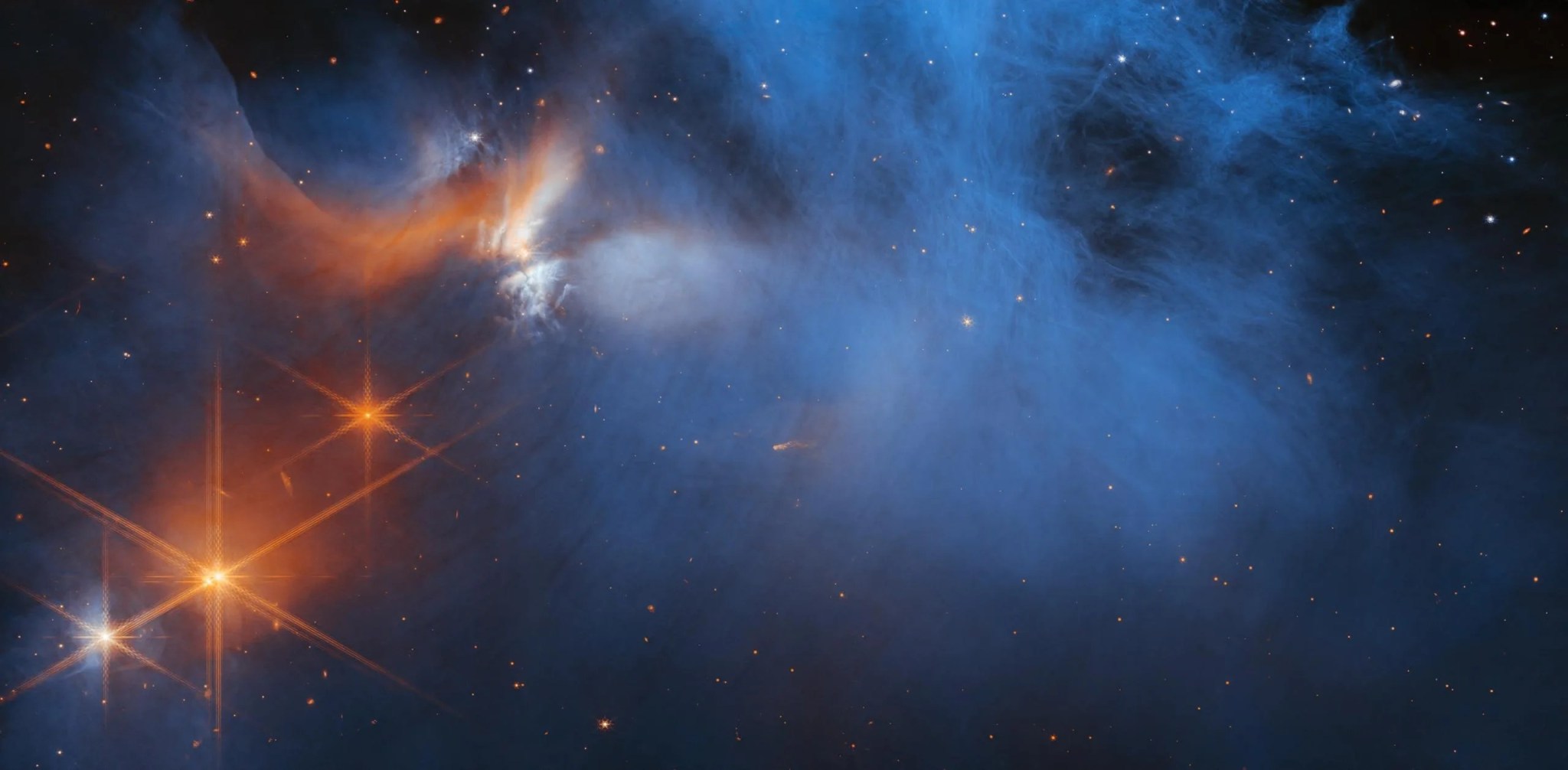 Imagen captada por Webb de la región central de la nube molecular oscura Camaleón I, que se encuentra a 630 años luz de distancia. Esta imagen muestra principalmente volutas de humo azul sobre un fondo oscuro. En la parte superior izquierda se aprecian además volutas naranjas y blancas. Justo debajo hay cuatro puntos de luz brillantes. Tres son de color naranja y uno es una mezcla de blanco y naranja. Cada uno de estos puntos tiene los característicos picos de difracción de ocho puntos de Webb.