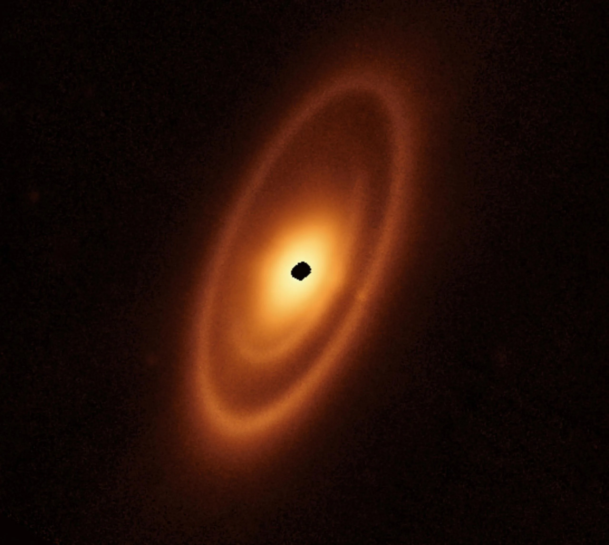 Un óvalo naranja se extiende desde la posición de la 1 en punto hasta la de las 7 horas. Presenta un anillo exterior prominente, un hueco más oscuro, un anillo intermedio, un hueco oscuro más estrecho y un disco interior brillante. En el centro hay una mancha negra irregular que indica la falta de datos.