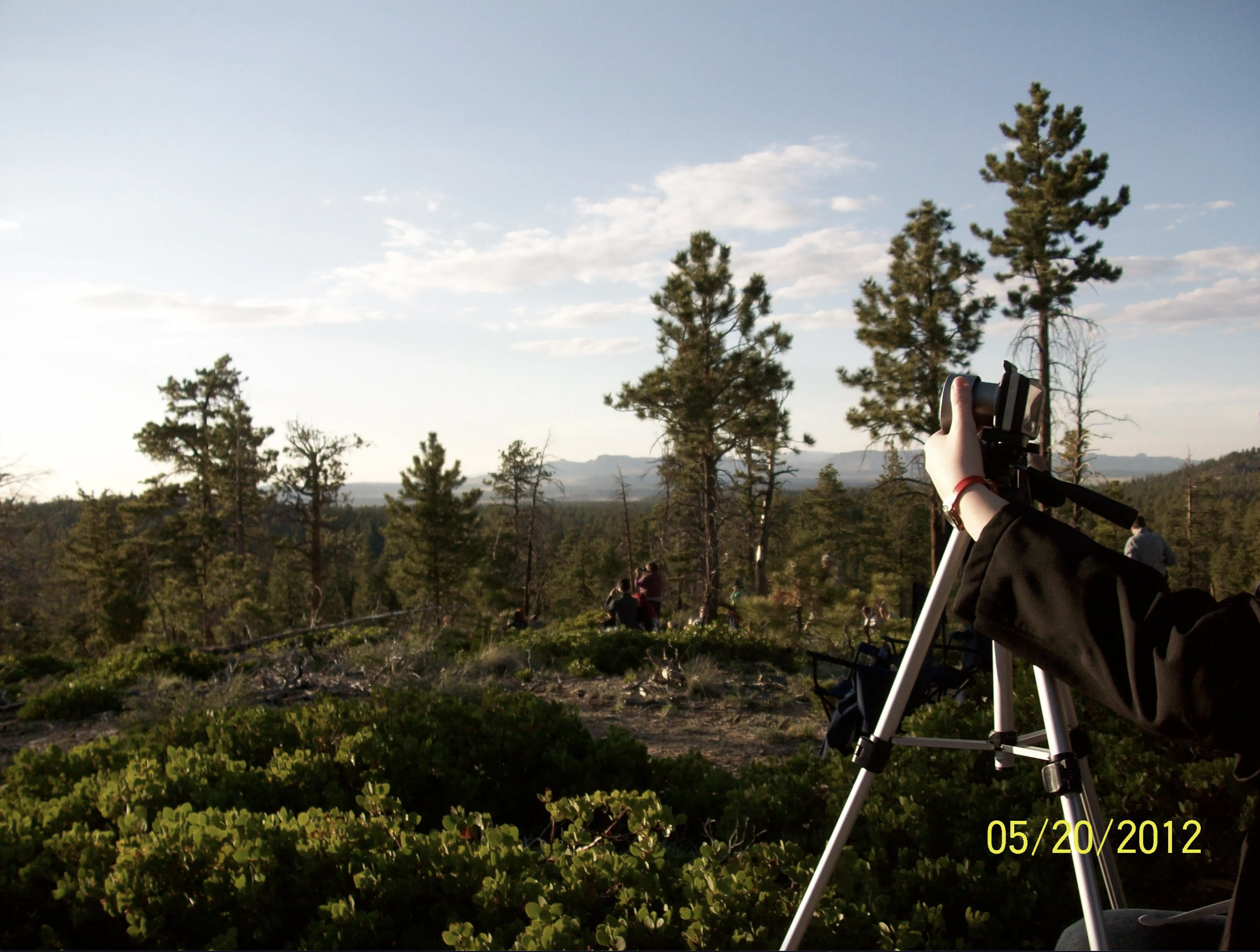 Esta imagen muestra un paisaje con árboles de fondo y un cielo celeste con algunas nubes blancas. A la derecha de la foto, se ve una cámara sobre un trípode, y una mano sujetándola.