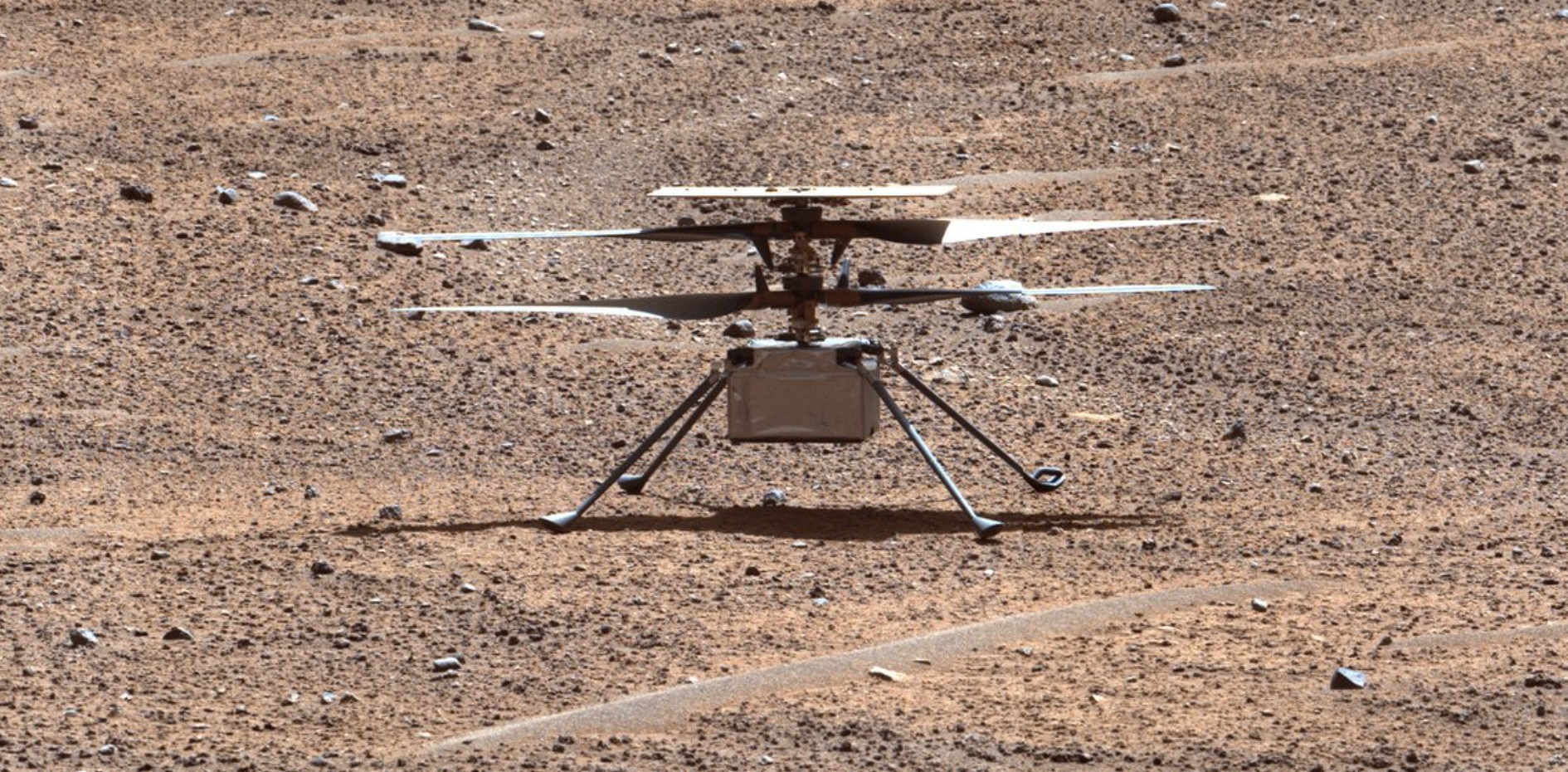 Imagen en color mejorado del helicóptero Ingenuity posado sobre la superficie rojiza marciana. El pequeño helicóptero tiene cuatro patas delgadas y cuatro aspas.