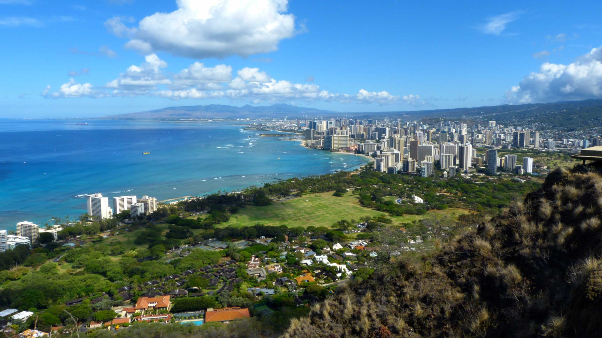 La imagen muestra una panorámica de un paisaje de Hawái, en el que se ve vegetación, edificaciones y mar. El cielo es celeste y aparecen algunas nubes blancas.