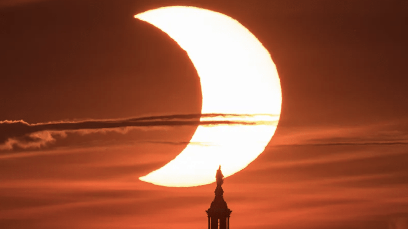 Eclipse solar parcial visto desde el edificio del Capitolio de Estados Unidos. El Sol aparece en forma de medialuna arriba y al centro de la foto. La bóbeda del Capitolio aparece como una silueta negra abajo y al centro. El cielo aparece en tonos de naranja intenso.