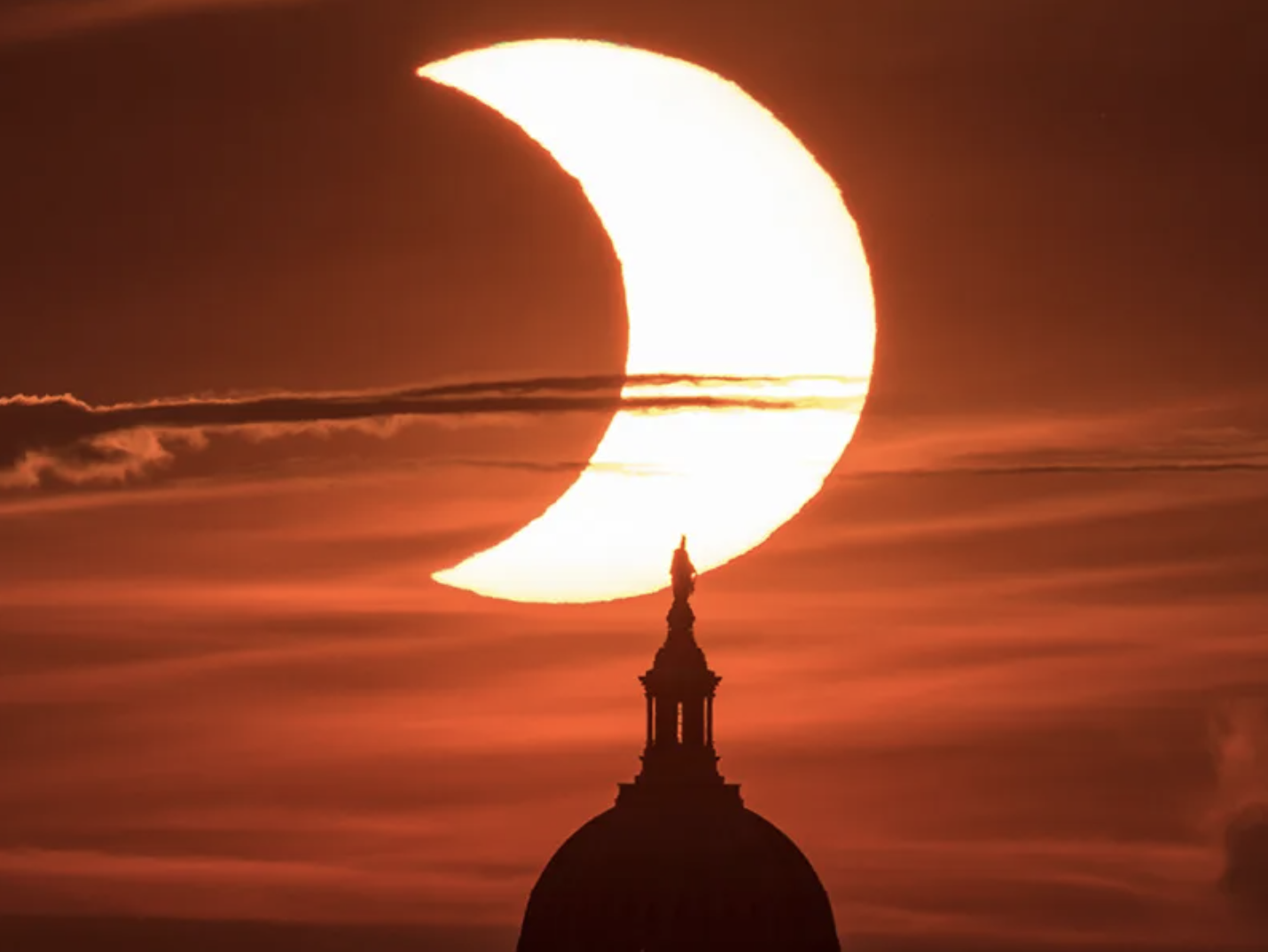 Eclipse solar parcial visto desde el edificio del Capitolio de Estados Unidos. El Sol aparece en forma de medialuna arriba y al centro de la foto. La bóbeda del Capitolio aparece como una silueta negra abajo y al centro. El cielo aparece en tonos de naranja intenso.