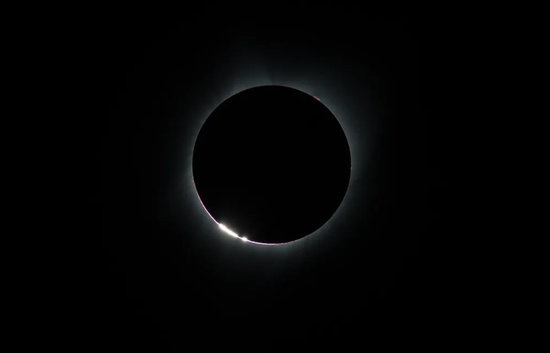 La imagen muestra un ejemplo de "perlas de Baily", las cuales aparecen a medida que la Luna hace su último tránsito delante del Sol durante un eclipse solar total. El Sol aparece en el centro de la imagen totalmente cubierto por la Luna, pero se ve un contorno brillante, más notable hacia la parte inferior izquierda del eclipse. El fondo es negro.