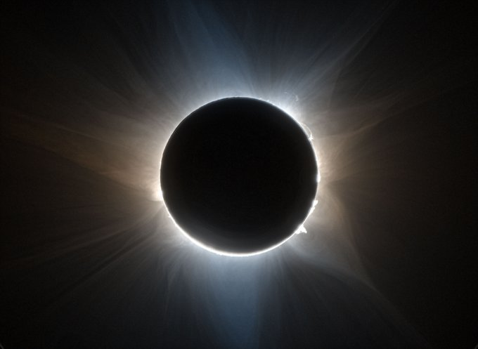 La sombra de la Luna oculta el Sol, que aparece como un disco negro. La corona solar se acentúa y las prominencias solares son visibles saltando desde la superficie. Del Sol emanan dramáticos susurros de energía solar. El resto de la imagen es oscuro.