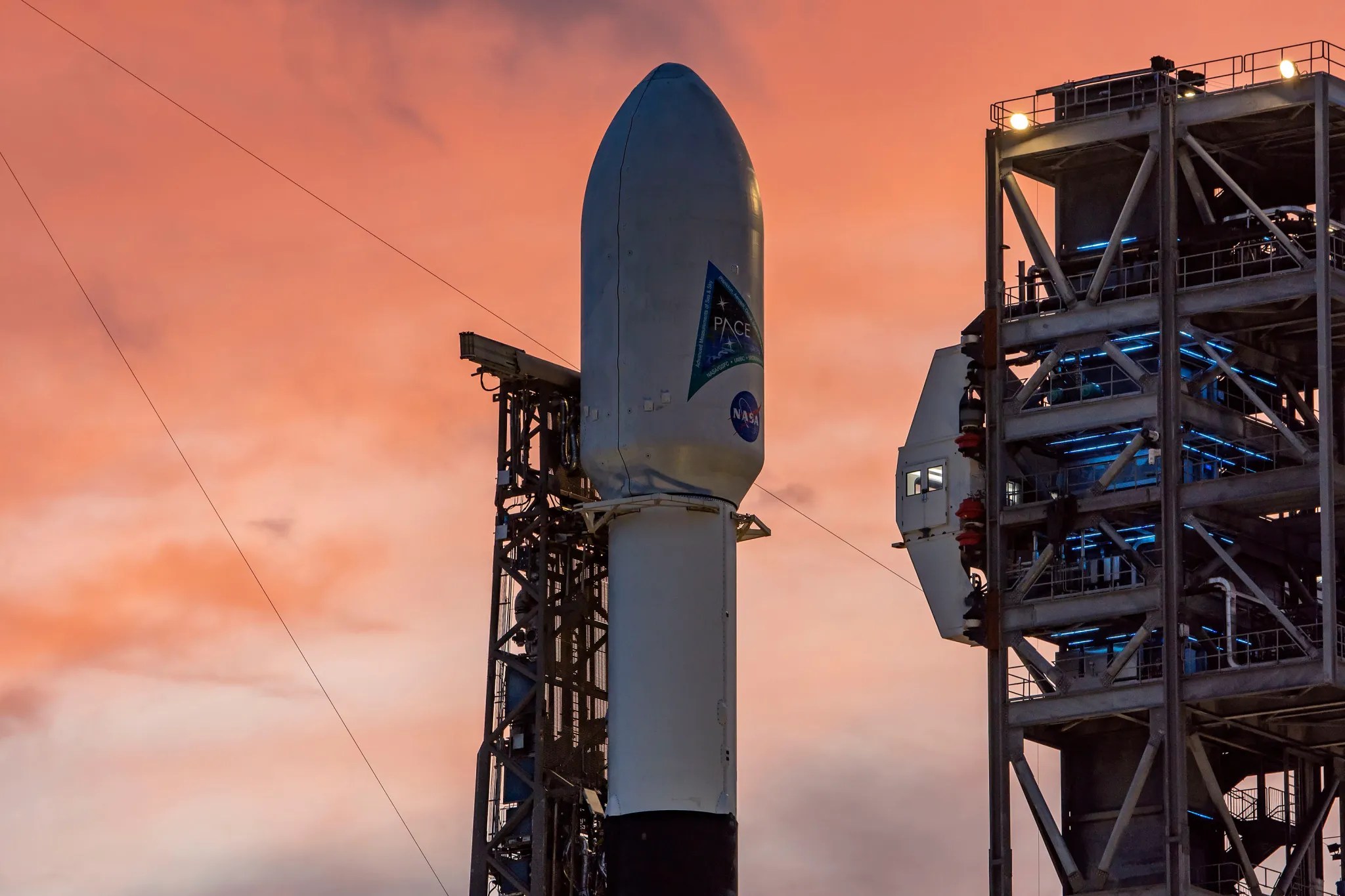 Una imagen de un cohete Falcon 9 en la plataforma de lanzamiento, con la parte superior del cohete visible en el centro de la imagen, junto a una plataforma de lanzamiento a la derecha.