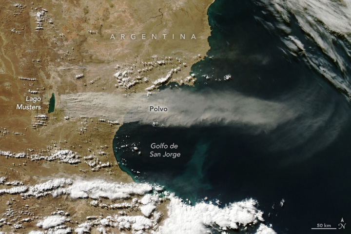 Imagen satelital de polvo saliendo del lago Colhué Huapi. El Golfo San Jorge, a la derecha, es de un azul intenso. Una costa curvilínea atraviesa el centro de la imagen de arriba abajo. A la izquierda, se ve un paisaje marrón arenoso. Una corriente de polvo gris claro comienza sobre el paisaje marrón y se extiende sobre el agua azul.